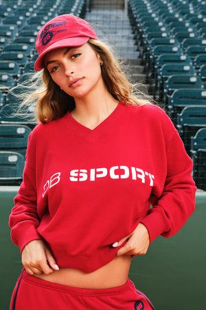 A woman wearing a red cropped V-neck sweatshirt with 'DB SPORT' printed in white across the chest, matching red sweatpants, and a red baseball cap, posed in front of stadium seating.
|M1