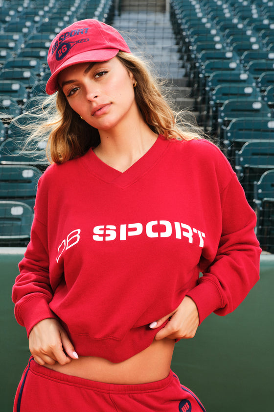A woman wearing a red cropped V-neck sweatshirt with 'DB SPORT' printed in white across the chest, matching red sweatpants, and a red baseball cap, posed in front of stadium seating.
|M1