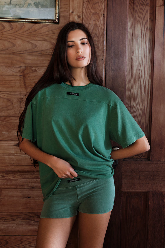Woman wearing a green t-shirt and shorts set against a wooden wall.
|M1