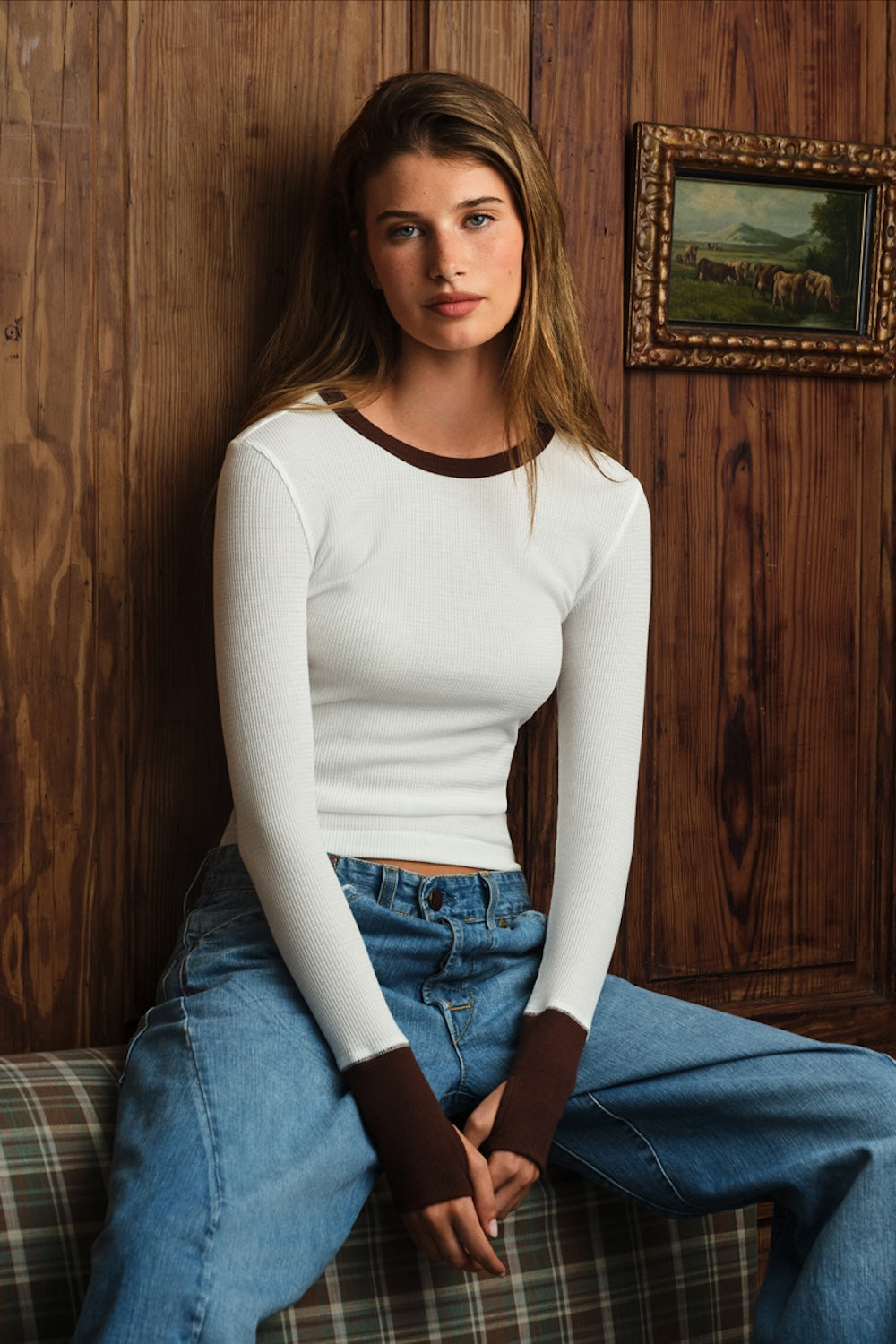 A woman sits on a plaid couch against a dark wood-paneled wall, wearing a fitted white long-sleeve top with contrasting brown trim at the crew neck and cuffs, paired with blue jeans.
|M1