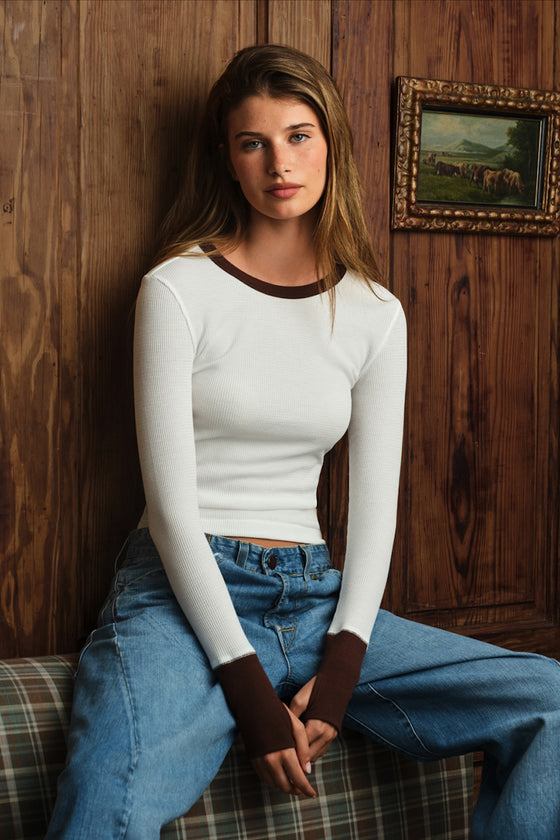 A woman sits on a plaid couch against a dark wood-paneled wall, wearing a fitted white long-sleeve top with contrasting brown trim at the crew neck and cuffs, paired with blue jeans.
|M1