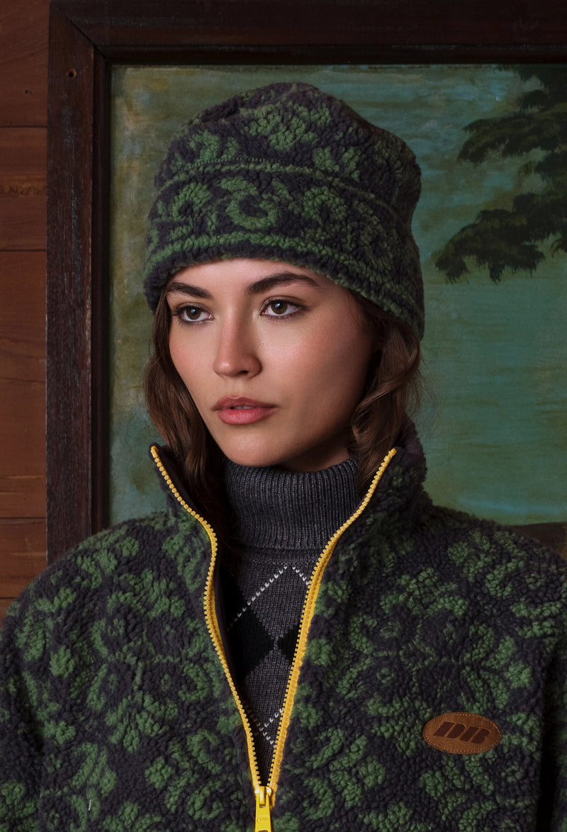 A woman wearing a dark green and black patterned knit beanie and matching zip-up fleece jacket with a yellow zipper, posed in front of a rustic wooden wall and framed painting.