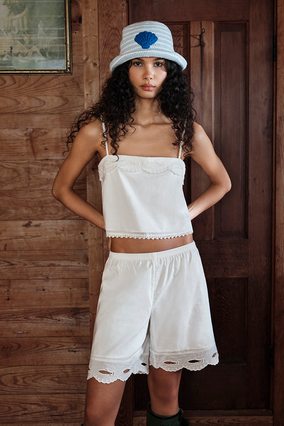 Woman wearing a white top and shorts with a blue flower hat in front of a wooden door.
|M1
