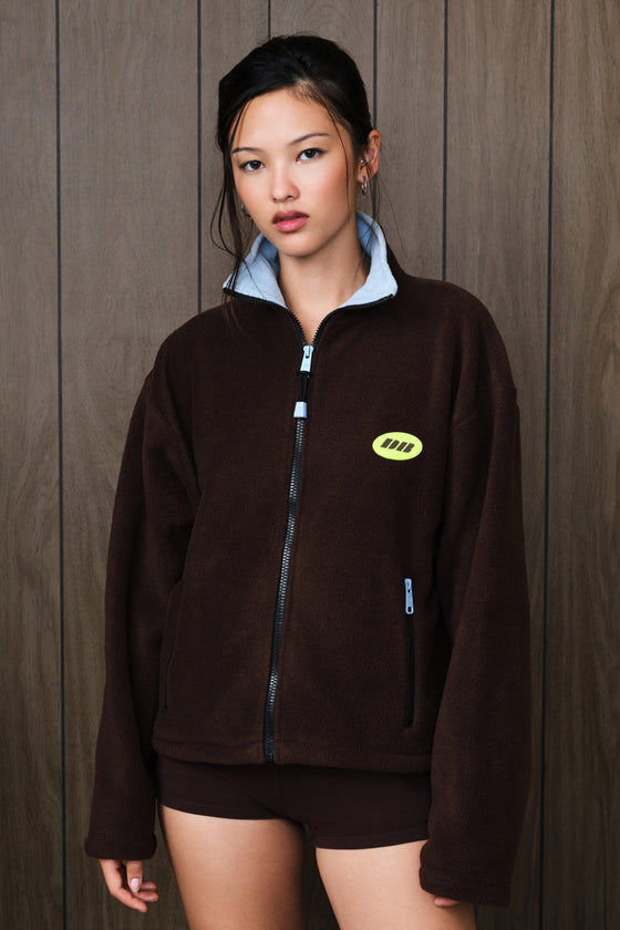 A model wearing a dark brown fleece zip-up jacket with a light blue inner collar and a small yellow-green logo patch on the chest, standing against a dark wood-paneled wall.
|M1