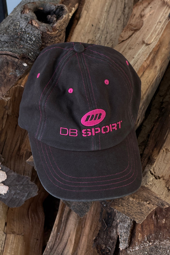A dark brown baseball cap with hot pink embroidery and stitching, featuring a 'DB SPORT' logo on the front, displayed on rustic wood logs.