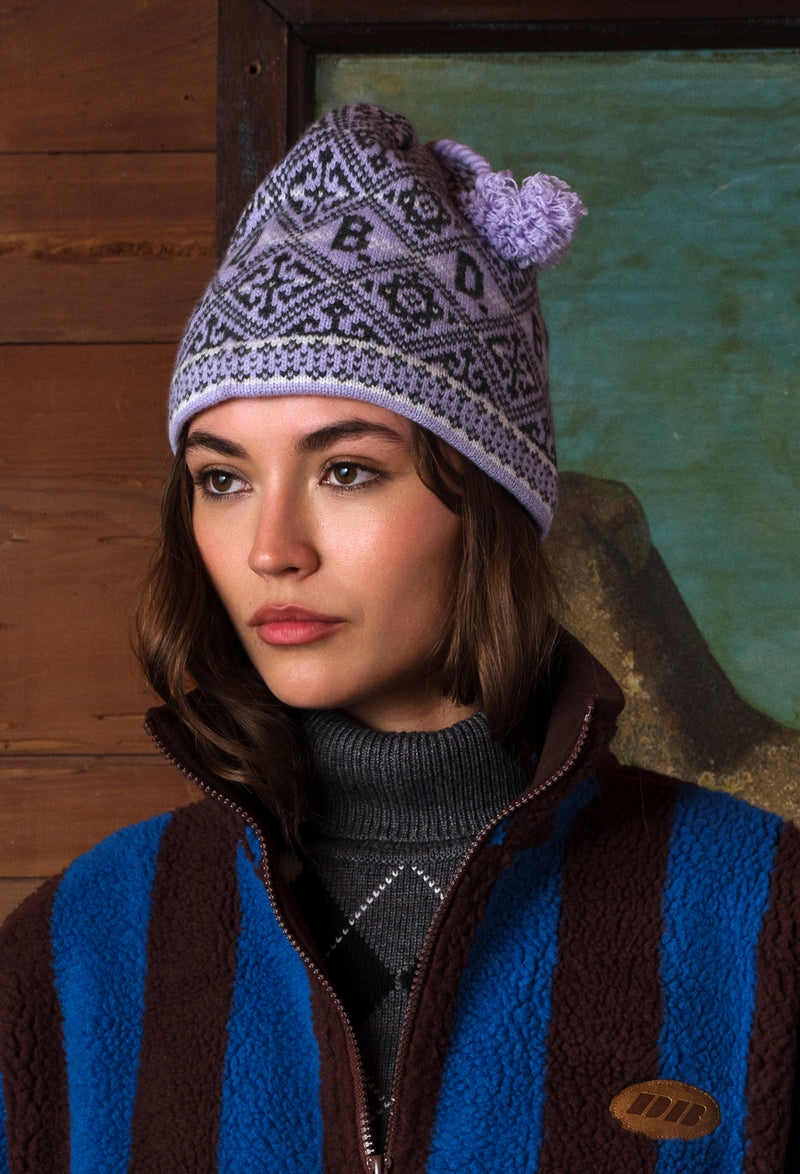 A woman wearing a purple and gray patterned knit beanie with a pom-pom, paired with a brown and blue striped fleece jacket over a gray turtleneck, set against a rustic wooden and green background.
