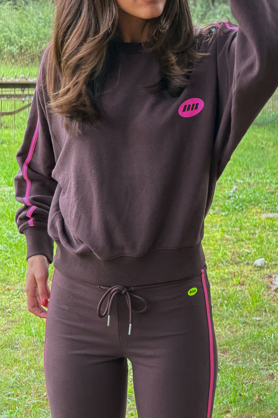 A woman wearing a matching dark brown sweatshirt and jogger pants set with bright pink side stripes and a small pink logo on the chest, photographed outdoors on grass.
|M1