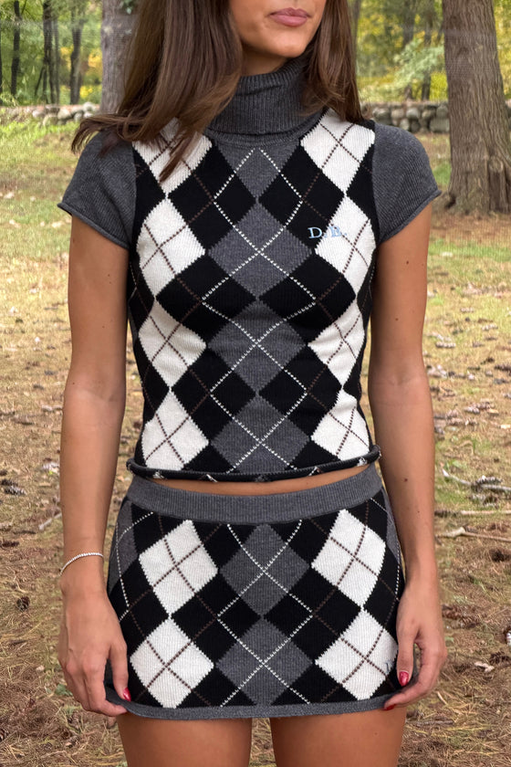 A woman wearing a matching knit two-piece set featuring a cropped turtleneck top and a high-waisted mini skirt, both in a black, white, and gray argyle pattern with solid gray short sleeves and waistband, photographed outdoors in a wooded setting.
|M1
