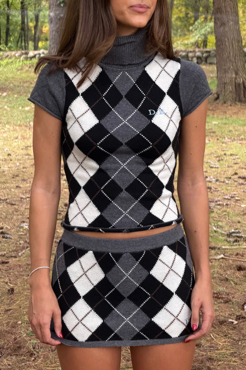A woman wearing a matching knit two-piece set featuring a cropped turtleneck top and a high-waisted mini skirt, both in a black, white, and gray argyle pattern with solid gray short sleeves and waistband, photographed outdoors in a wooded setting.
|M1