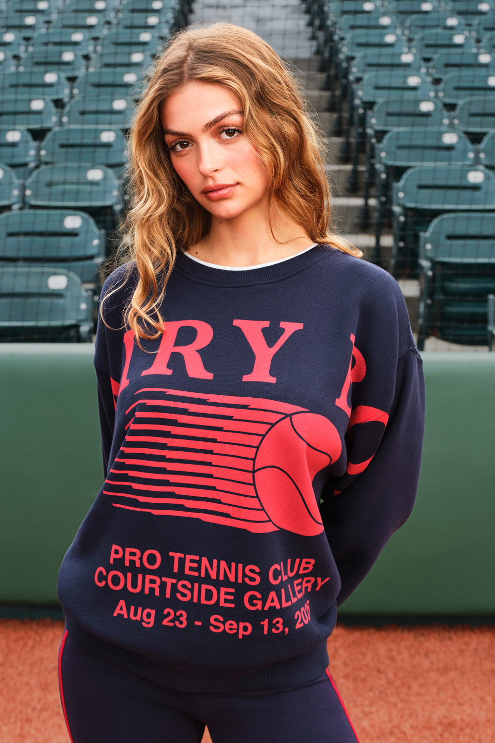 A woman stands on a tennis court wearing a navy blue sweatshirt with a red graphic of a tennis ball and the text 'PRO TENNIS CLUB COURTSIDE GALLERY' and 'Aug 23 - Sep 13, 2025'.
|M1