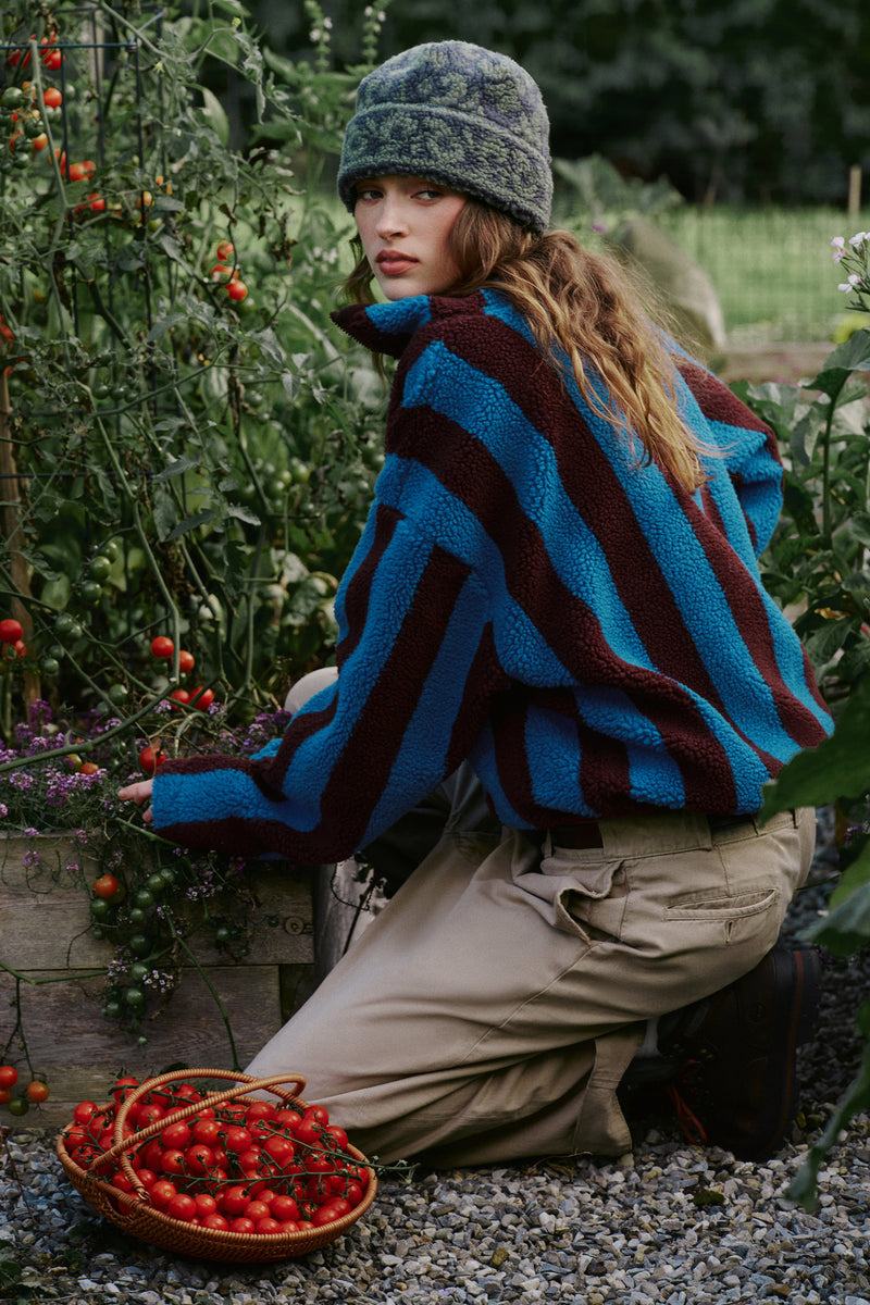 A woman kneels in a garden wearing a blue and dark red striped fleece jacket, beige pants, and a green knit beanie, with a basket of red tomatoes on the ground nearby.
|M1