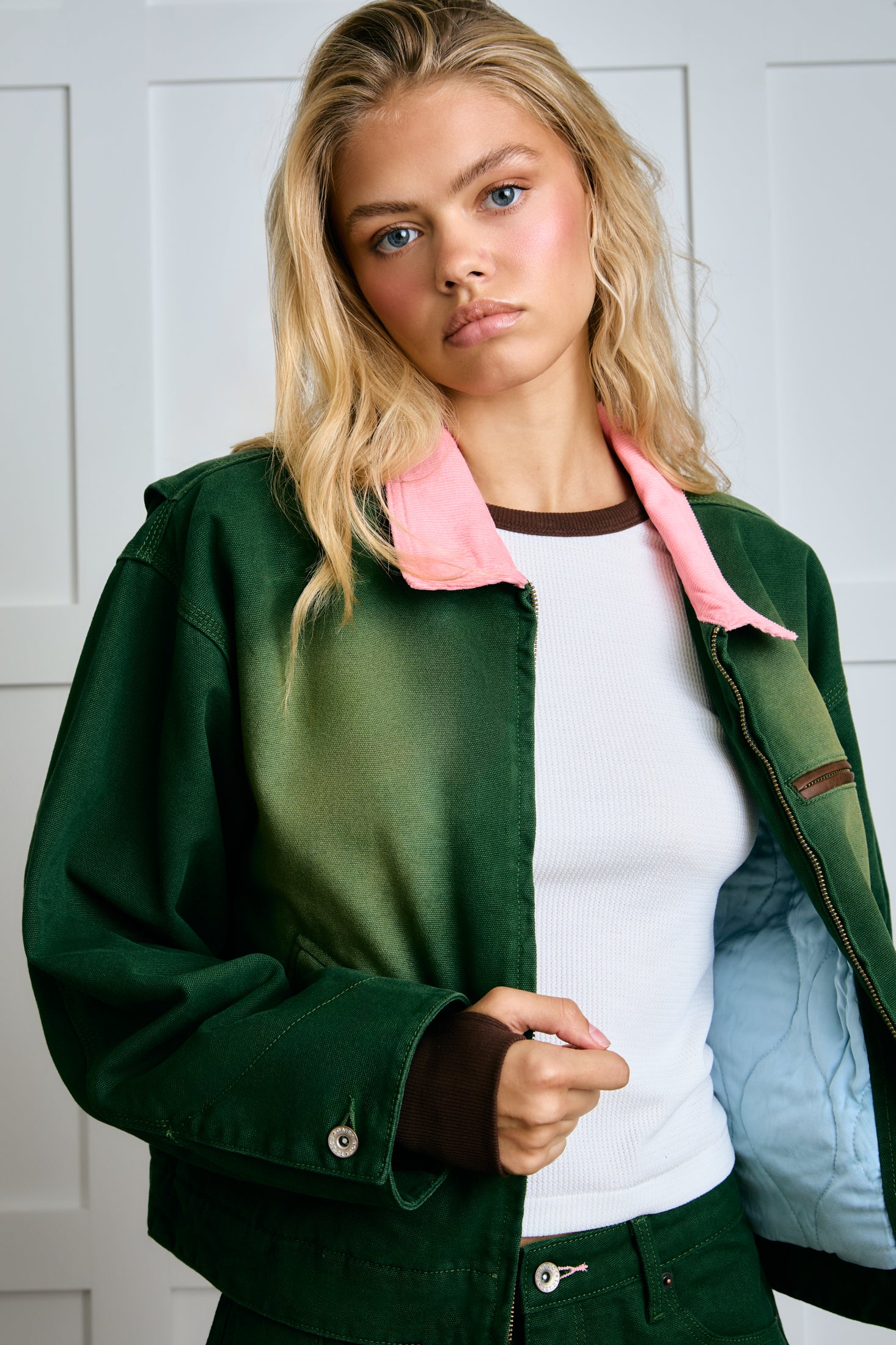 Woman wearing a green jacket with a pink collar over a white shirt, standing against a white wall. |m1