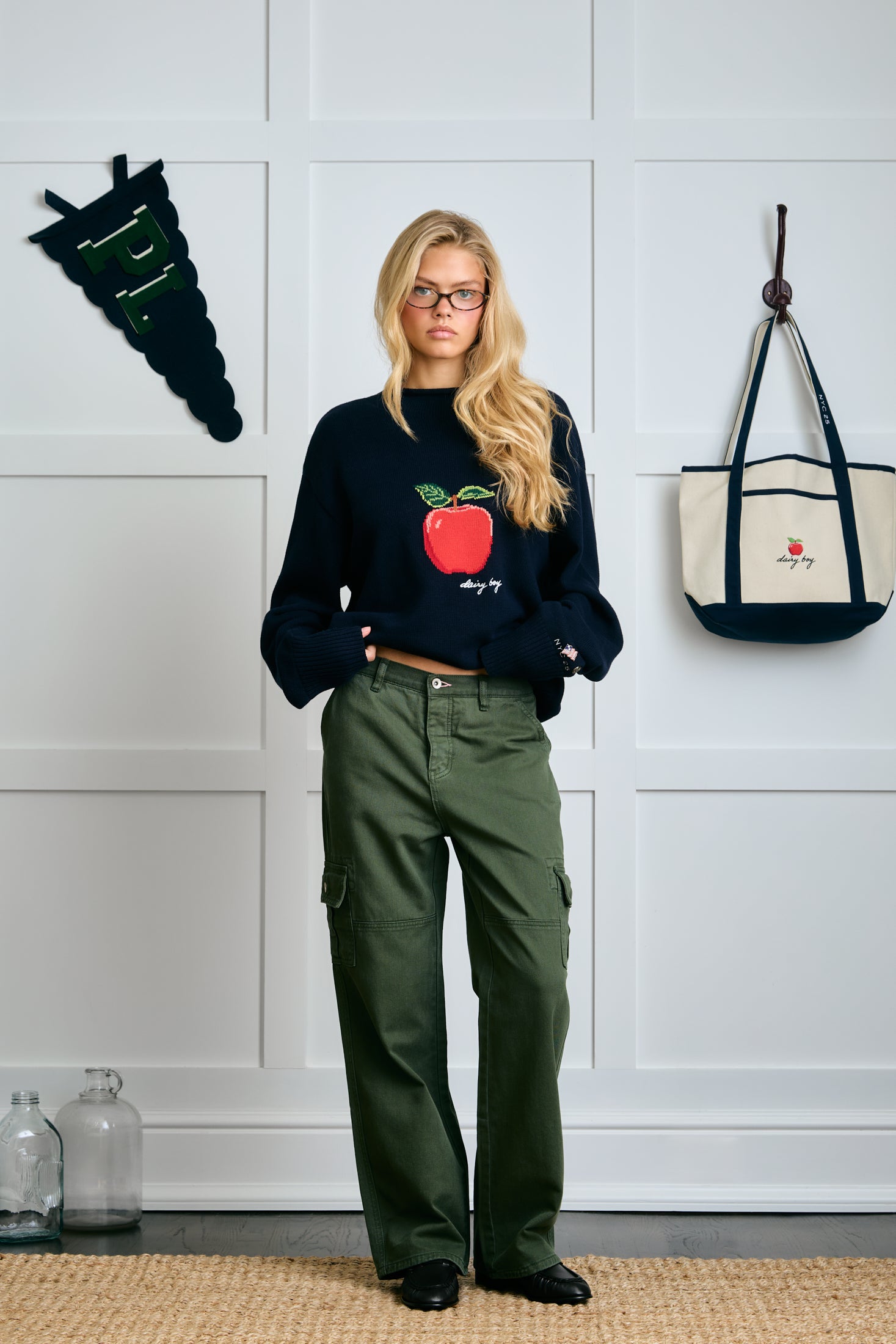 Person wearing a black sweater with a red apple design and green pants standing in front of a white paneled wall. |m1
