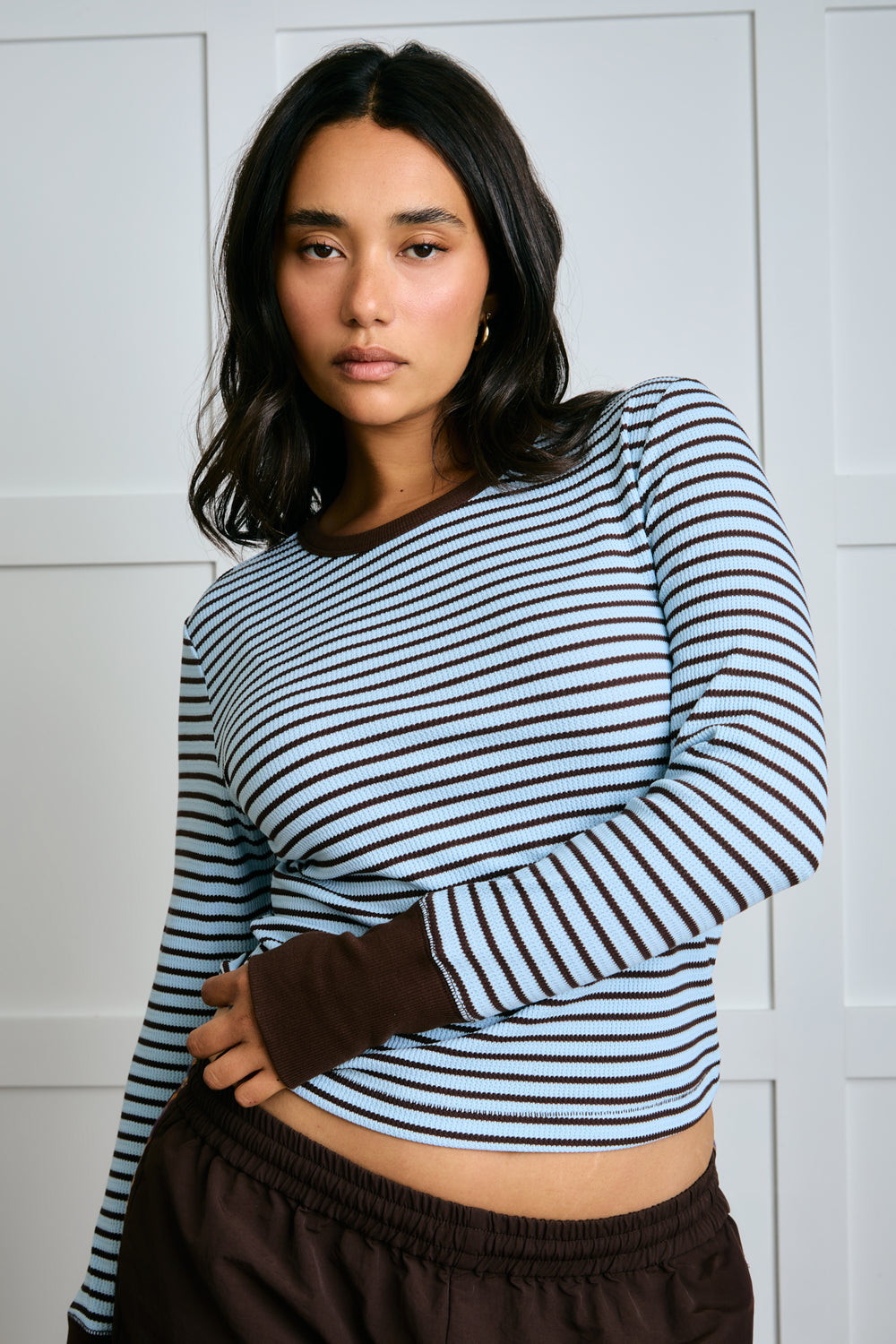 Woman wearing a striped long-sleeve top and brown pants against a white paneled wall. |m1
