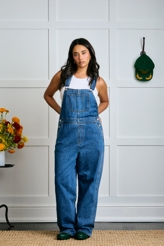 Person wearing blue denim overalls standing in a room with a white paneled wall. |m1
