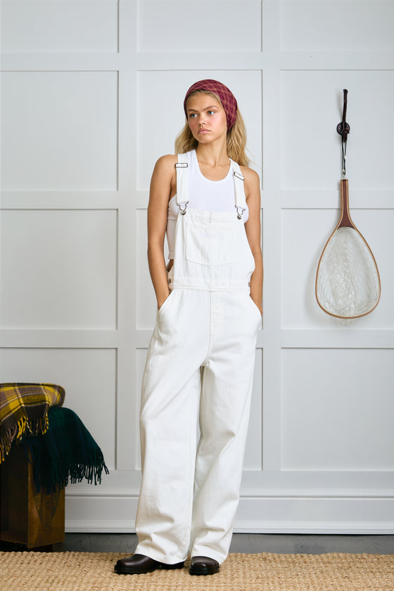 Woman wearing white overalls standing in a room with a white wall and a fishing net hanging on it. |m1