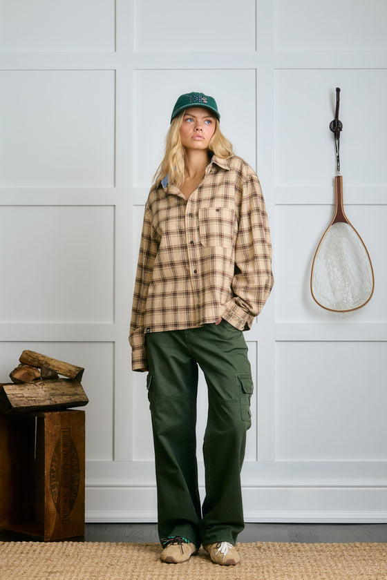 Person wearing a plaid shirt and green pants standing in a room with a white paneled wall. |m1