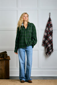 Woman wearing a green plaid shirt and blue jeans standing in a room with a plaid blanket and wooden chest. |m1