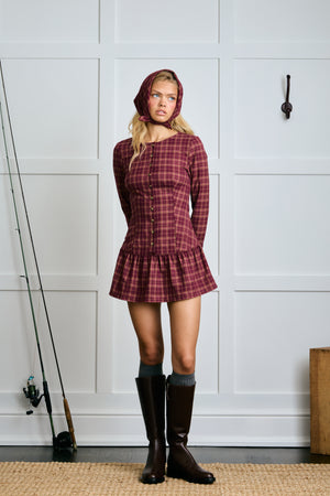 Woman wearing a plaid dress and knee-high boots standing in a room with white paneled walls.  |m1