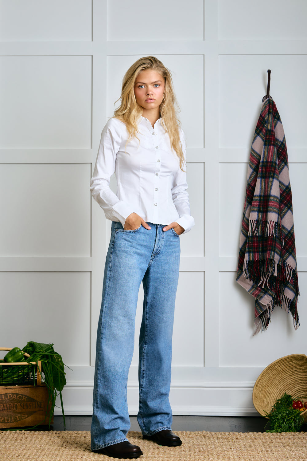 Woman wearing a white shirt and blue jeans standing against a white paneled wall with a plaid blanket. |m1