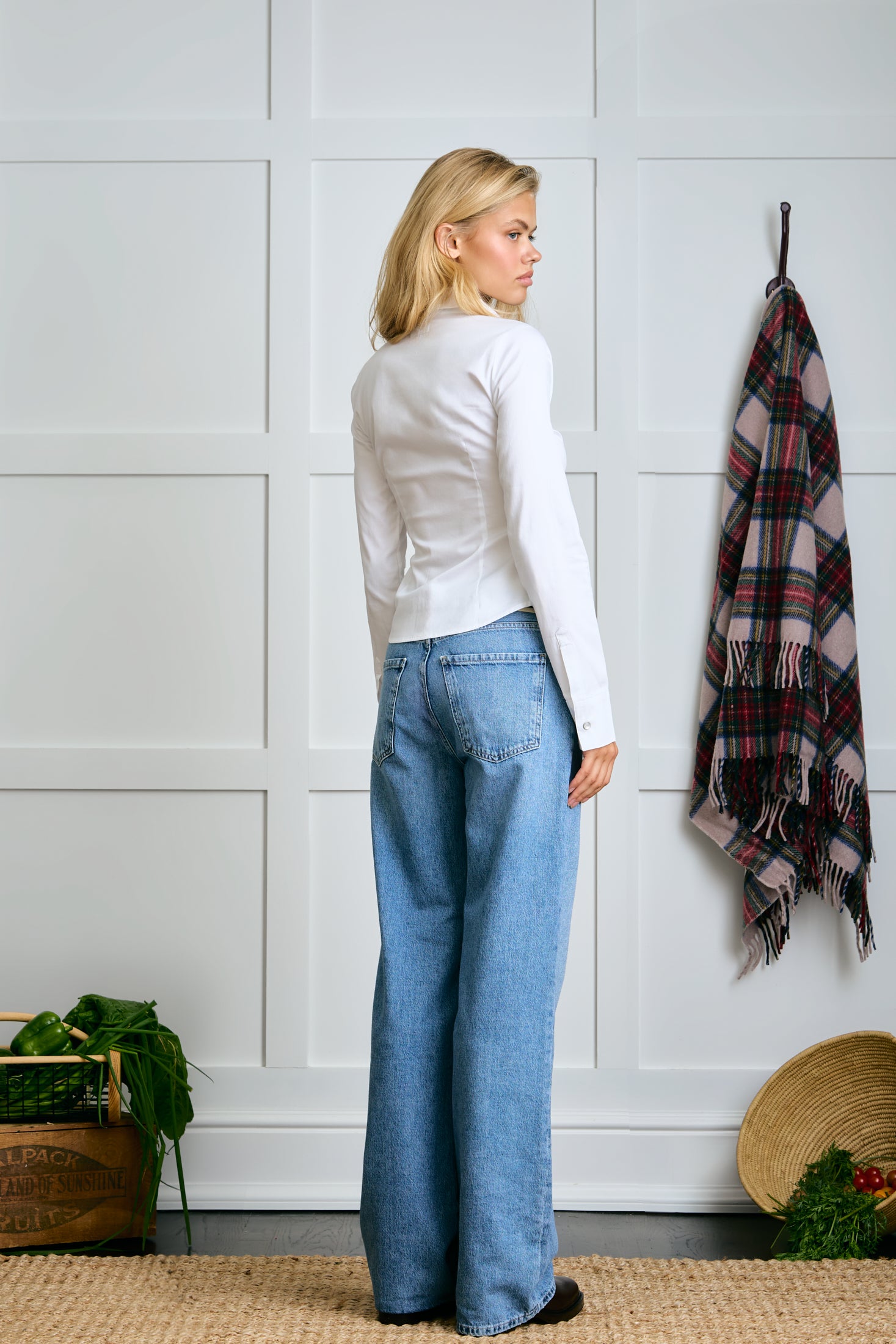 Woman wearing a white sweater and blue jeans standing against a white paneled wall with a plaid blanket and plant in the background.  |m1