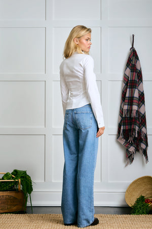 Woman wearing a white sweater and blue jeans standing against a white paneled wall with a plaid blanket and plant in the background.  |m1