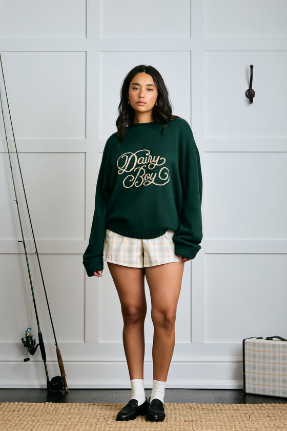 Person wearing a green sweatshirt with text, standing in a room with a white paneled wall. |m1