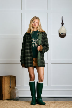 Woman wearing a plaid shirt over a green sweater with text, shorts, and green boots in a room with a wooden box and hat on a hook. |m1