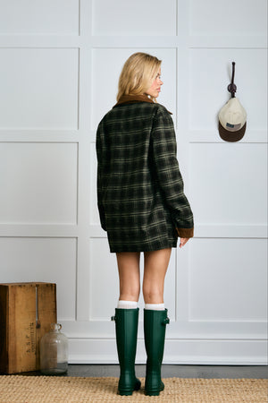 Person wearing a plaid coat and green rain boots standing in front of a white paneled wall. |m1