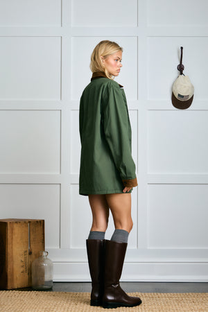 Woman wearing a green coat and brown boots standing against a white paneled wall.|m1