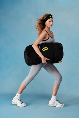 Woman holding a black duffel bag with a yellow logo against a blue background  |M1