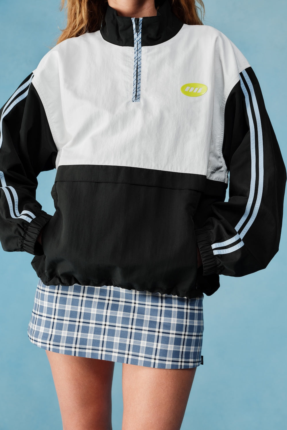 Person wearing a black and white jacket with a logo and plaid skirt on a blue background |M1