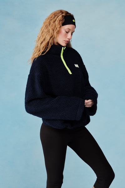 Woman wearing a navy fleece pullover with a green zipper on a light blue background  |M1