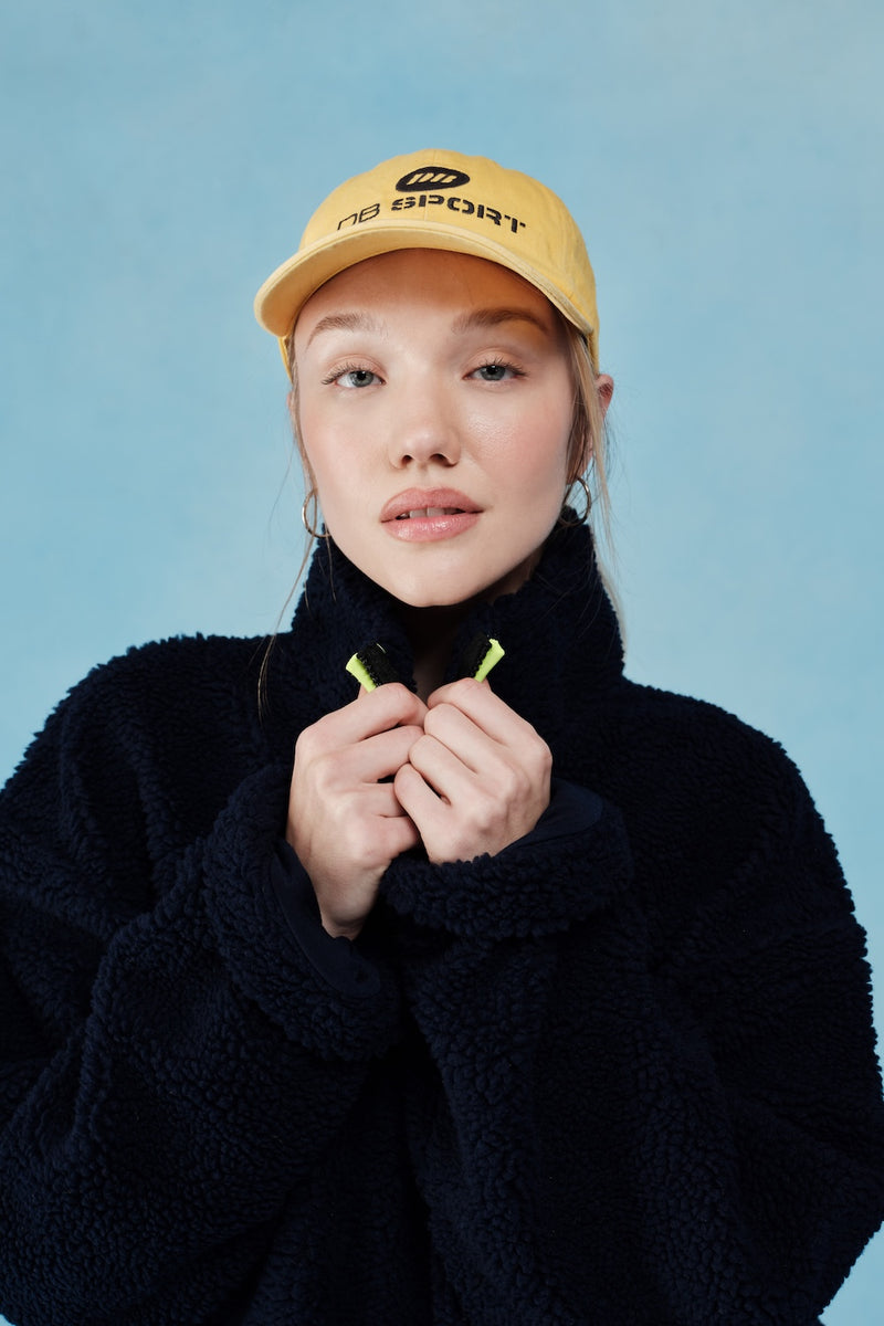 Person wearing a yellow cap with DB SPORT text and holding collar against a blue background