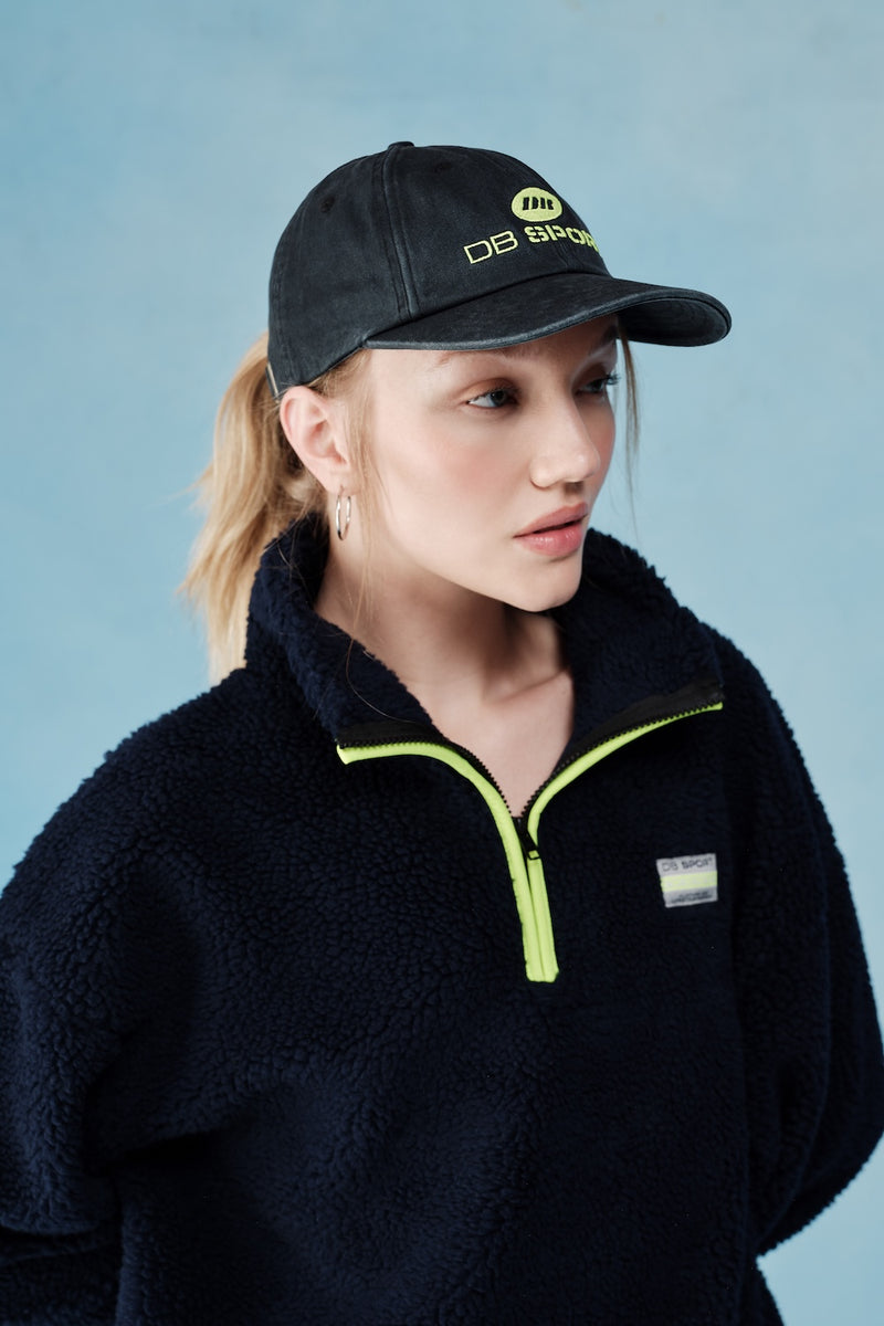 Person wearing a black cap and dark jacket with a visible brand logo against a light blue background