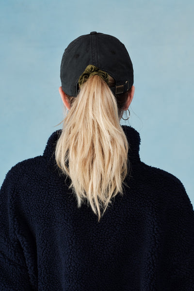 Person wearing a black cap and black jacket with blonde hair in a ponytail against a light blue background