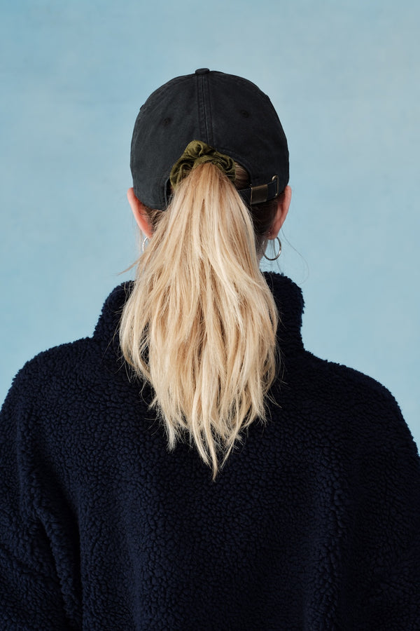 Person wearing a black cap and black jacket with blonde hair in a ponytail against a light blue background