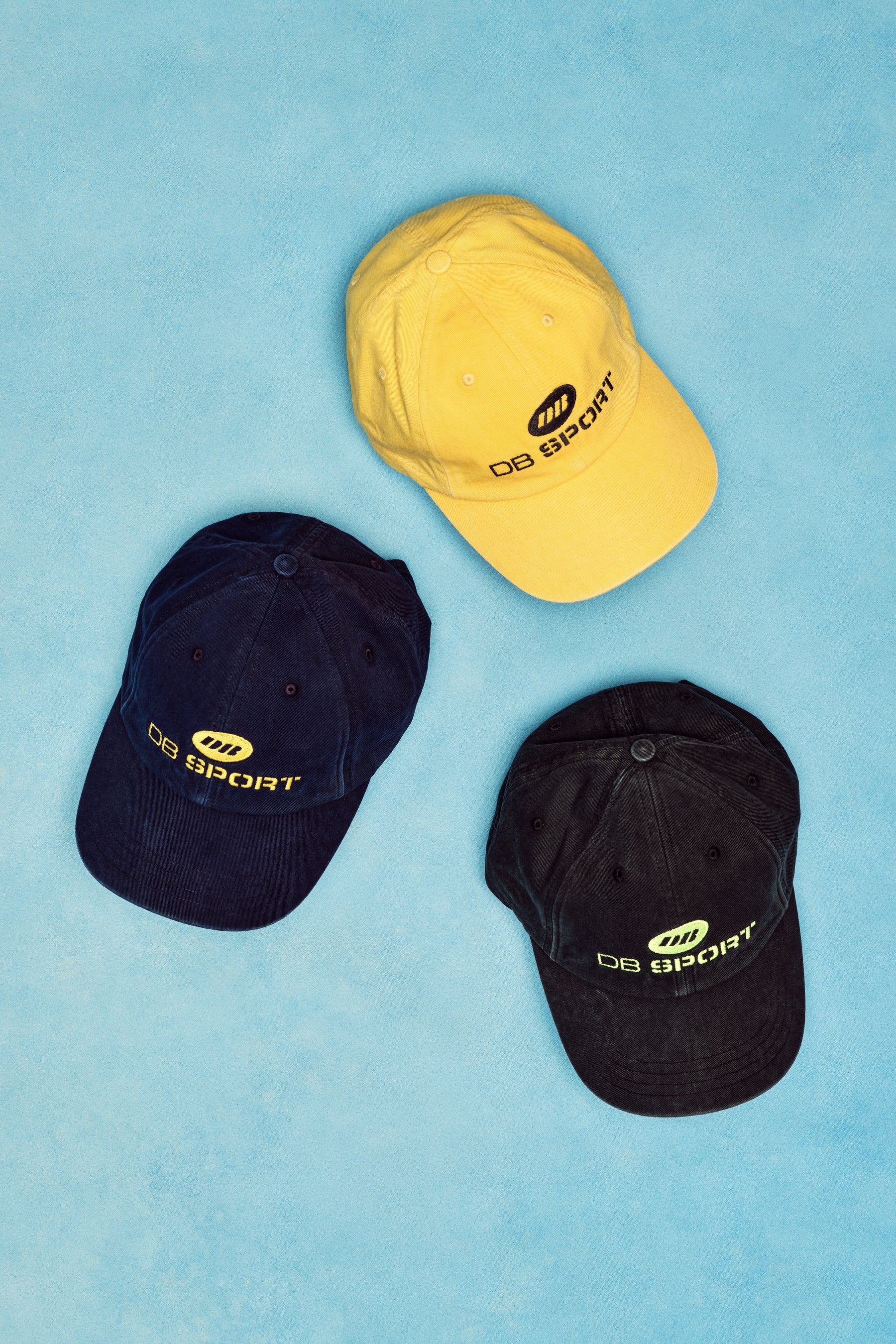 Three caps in yellow, black, and navy on a blue background with 'DB Sport' branding.