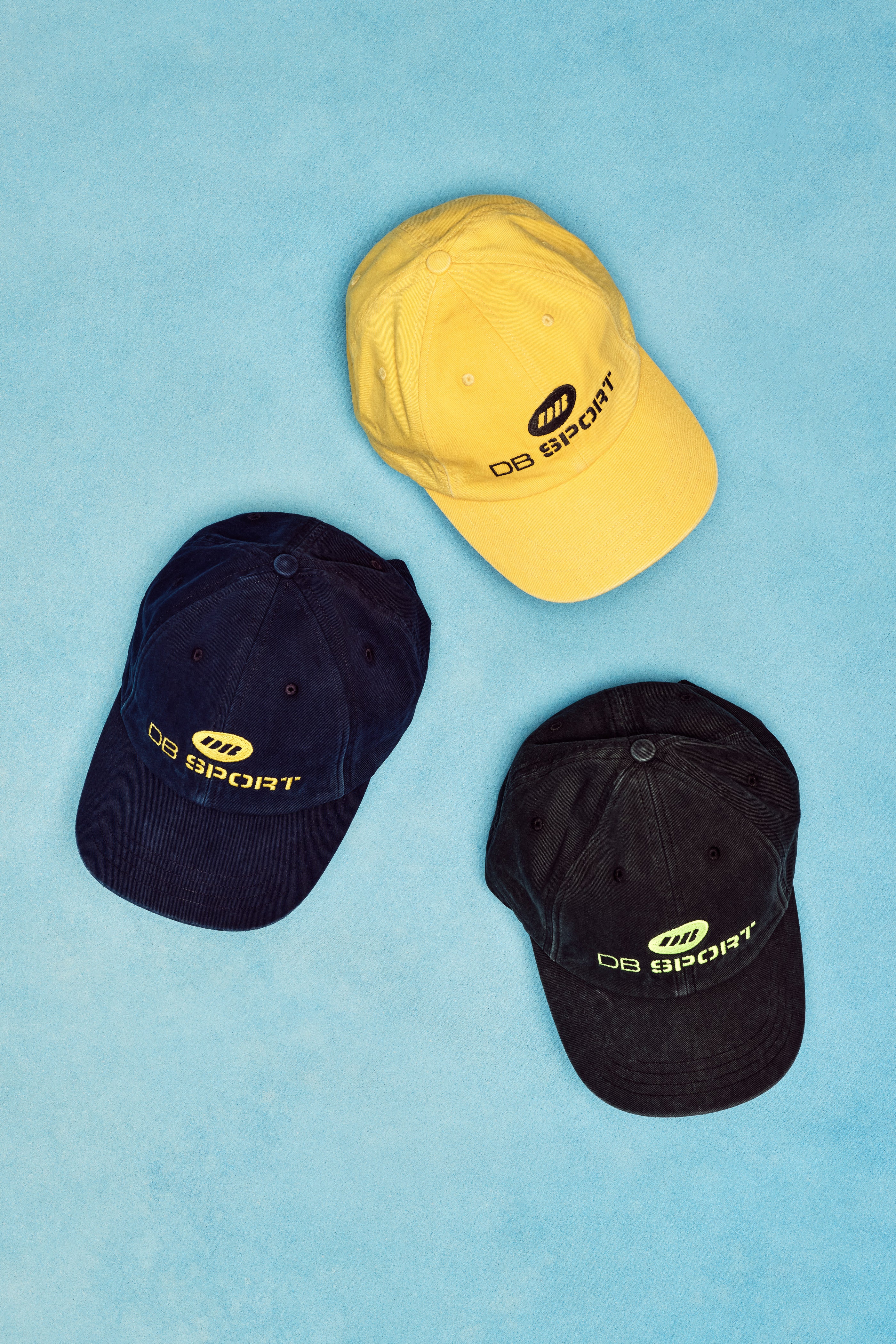 Three caps in yellow, black, and navy on a blue background with 'DB Sport' branding.