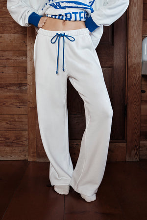 Person wearing a white sweatshirt with blue accents and white sweatpants against a wooden background
|M1