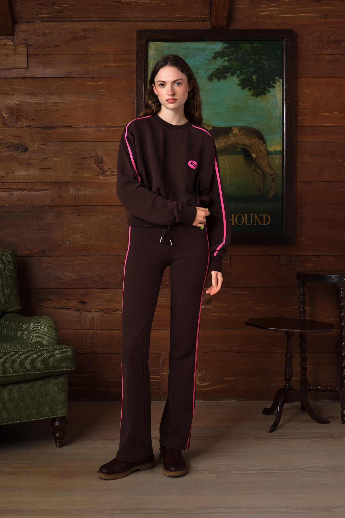 A woman stands in a rustic room with wood-paneled walls wearing a dark brown tracksuit with bright pink piping and a small pink logo on the chest. The set includes a long-sleeve top and matching pants.
|M2