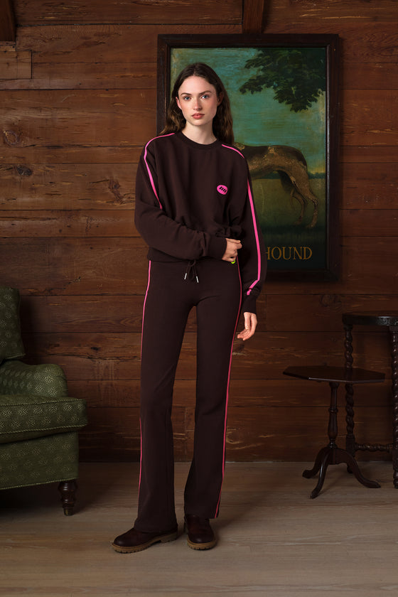 A woman stands in a rustic room with wood-paneled walls wearing a dark brown tracksuit with bright pink piping and a small pink logo on the chest. The set includes a long-sleeve top and matching pants.
|M2