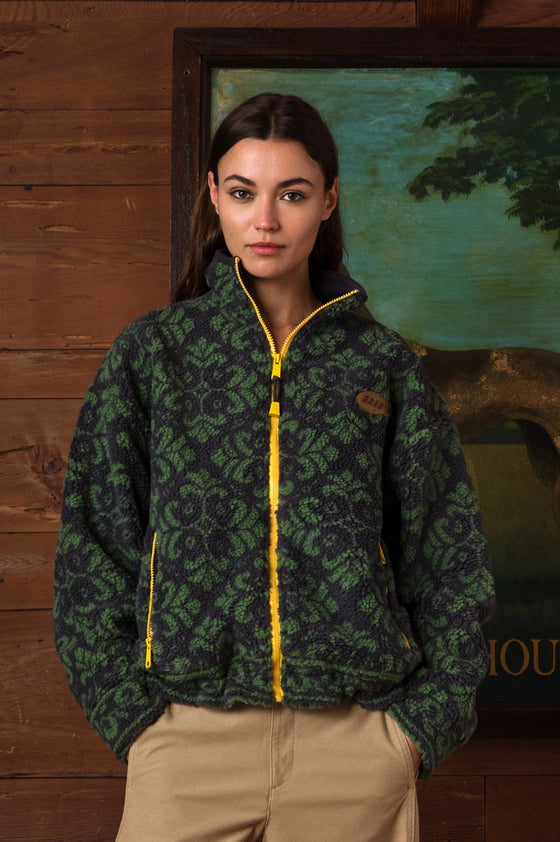 A woman wearing a dark green and black damask-patterned fleece jacket with bright yellow zipper accents, paired with beige pants, standing in front of a rustic wooden wall and a framed landscape painting.
|M2