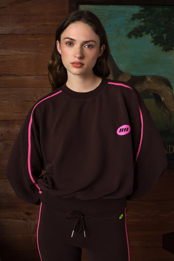 A woman wearing a dark brown sweatshirt with bright pink piping and a small pink chest logo, paired with matching brown pants with pink piping, set against a rustic wooden wall background.
|M2
