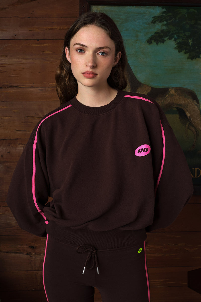 A woman wearing a dark brown sweatshirt with bright pink piping and a small pink chest logo, paired with matching brown pants with pink piping, set against a rustic wooden wall background.
|M2
