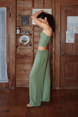 Woman in a green outfit standing in a room with wooden walls and a door.
|M1