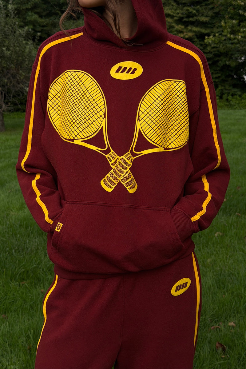 A person wearing a maroon and yellow sweatsuit outdoors on grass, with a large yellow tennis racket graphic across the hoodie and matching yellow piping along the sleeves and pants.
|M1
