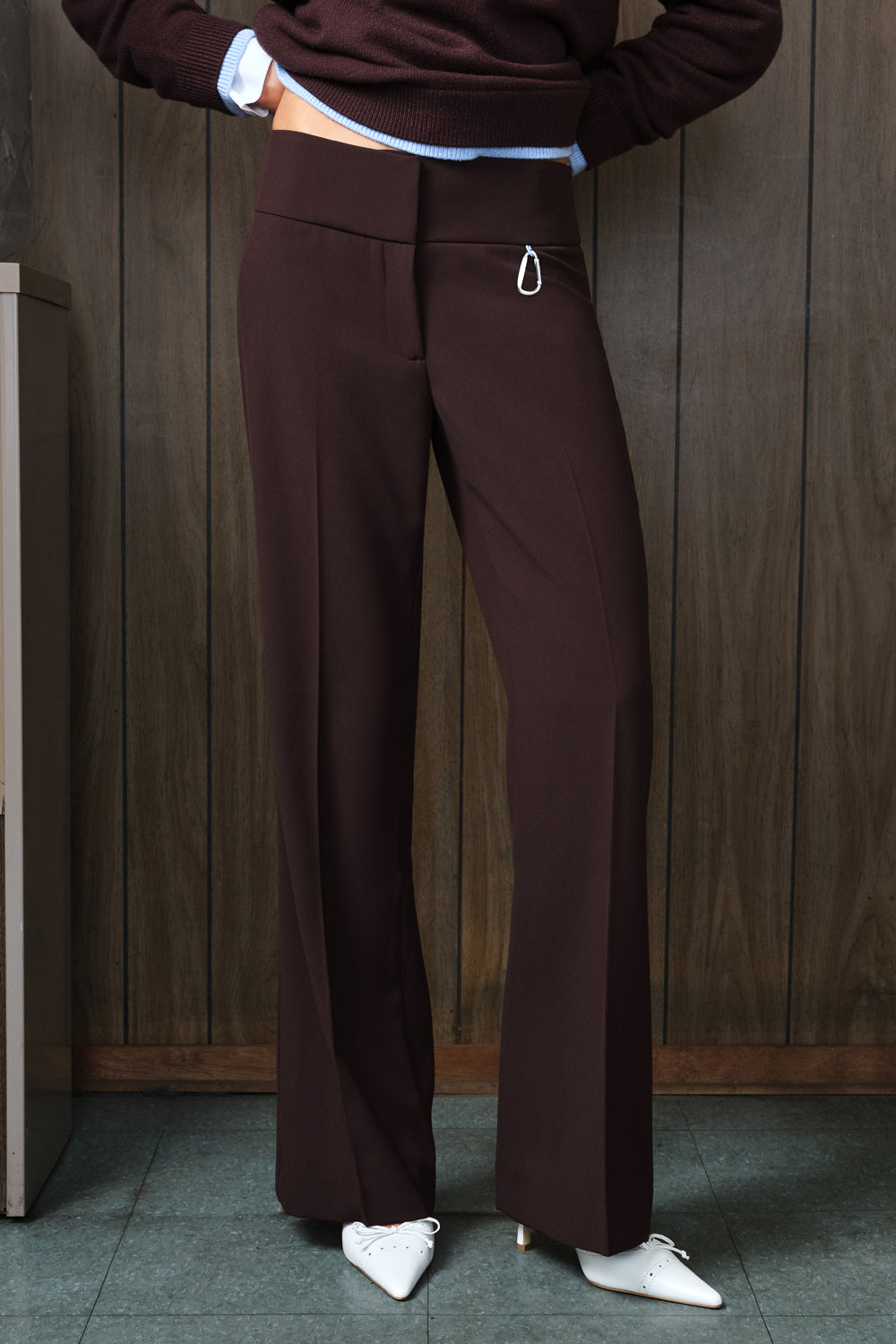 A person wearing dark brown wide-leg trousers with a small silver ring detail on the front pocket, paired with a dark brown sweater and white pointed shoes, standing against a dark wood-paneled wall.
|M1