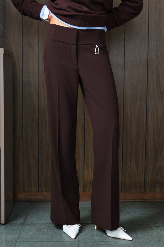 A person wearing dark brown wide-leg trousers with a small silver ring detail on the front pocket, paired with a dark brown sweater and white pointed shoes, standing against a dark wood-paneled wall.
|M1