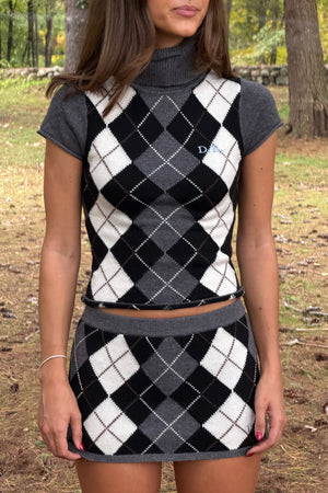 A woman wearing a matching knit two-piece set featuring a cropped turtleneck top and a mini skirt, both in a black, white, and gray argyle pattern with solid gray short sleeves and waistband, photographed outdoors in a wooded setting.
|M2